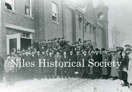 A large group of Niles citizens standing outside the fire station on Franklin Alley on a cold wintry day. The horse stalls have been converted to parking areas for the fire truck.